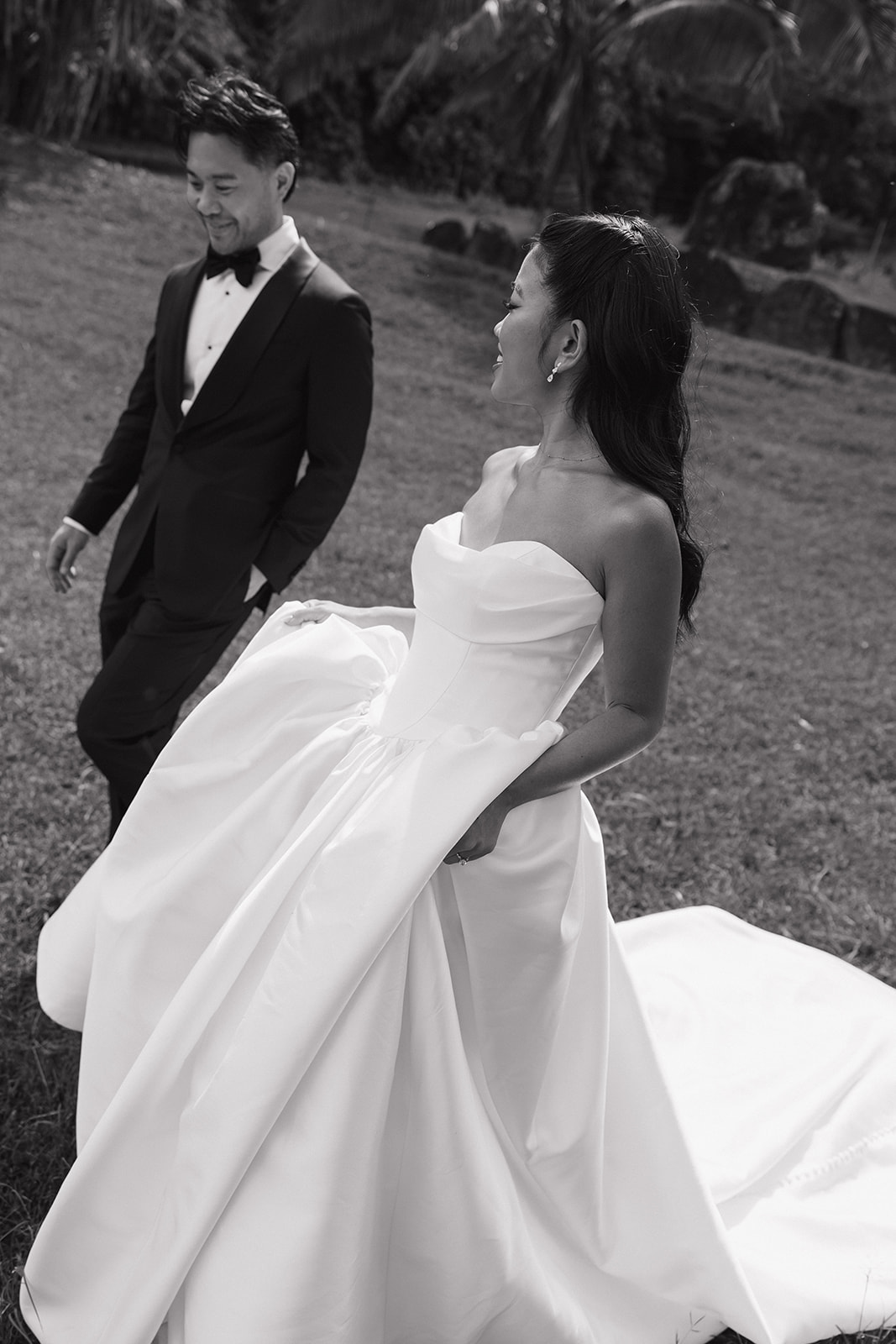 Black and white photo of bride lifting her ball gown skirt while laughing with her groom walking beside her on lush green grass