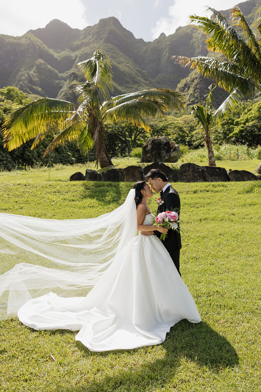 Bride and groom touching foreheads in an intimate embrace while her cathedral-length veil billows dramatically in the wind against lush palm trees and volcanic rocks