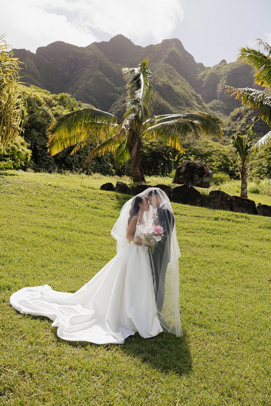 Couple sharing an intimate forehead-to-forehead moment under the bride's veil at a Kualoa Ranch Wedding, surrounded by palm trees and the Ko'olau mountains