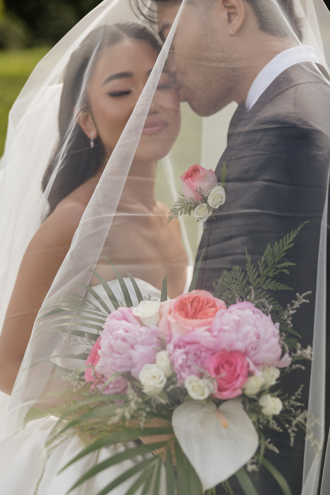 Romantic under-the-veil portrait at a Kualoa Ranch Wedding showing the bride's lush tropical bouquet of pink peonies, coral garden roses, white calla lilies, and palm fronds while the couple leans in close beneath the sheer fabric