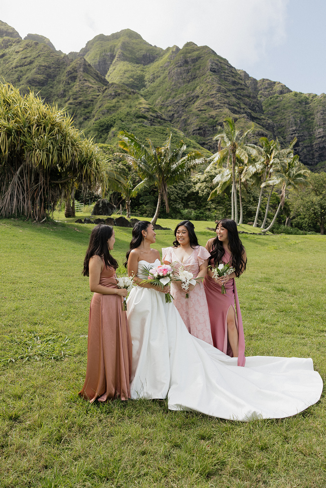 Bride with three bridesmaids in coordinating pink gowns gathered on the lawn holding tropical bouquets, with the dramatic green Ko'olau mountain range and swaying palm trees filling the entire background