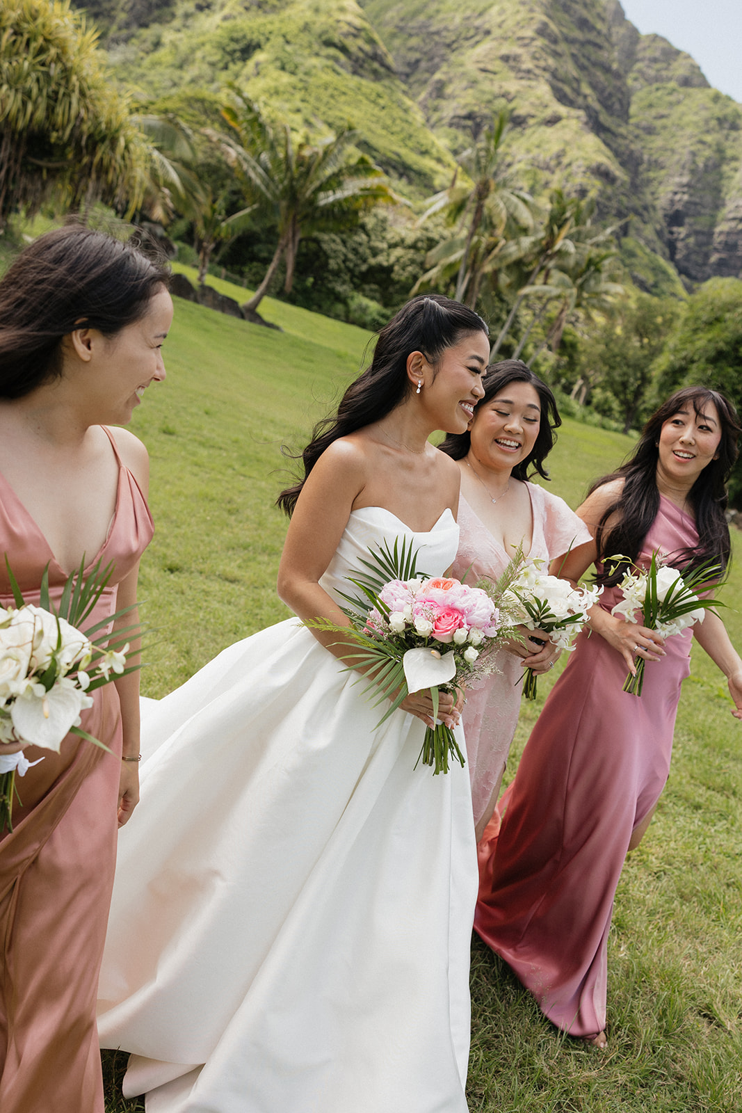 Bride and three bridesmaids in mismatched dusty rose and terracotta satin gowns laughing and walking together on the grass at their Kualoa Ranch Wedding, holding tropical bouquets with the lush Ko'olau cliffs behind them