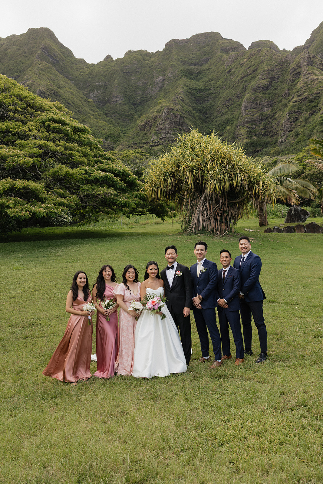 Wedding party group photo at a Kualoa Ranch Wedding with three bridesmaids in mismatched pink gowns standing alongside three groomsmen in navy suits with the stunning Ko'olau mountain range behind them
