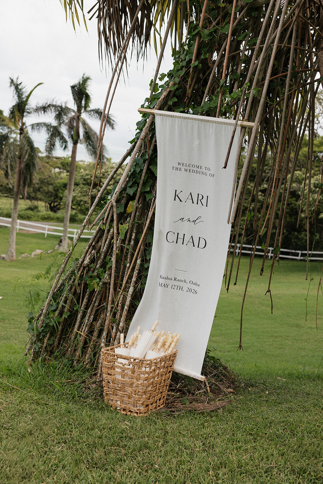 Welcome banner sign reading "Kari and Chad — Kualoa Ranch, Oahu, May 12th, 2026" hanging from a bamboo rod against a rustic driftwood arch with a woven basket of parasols below
