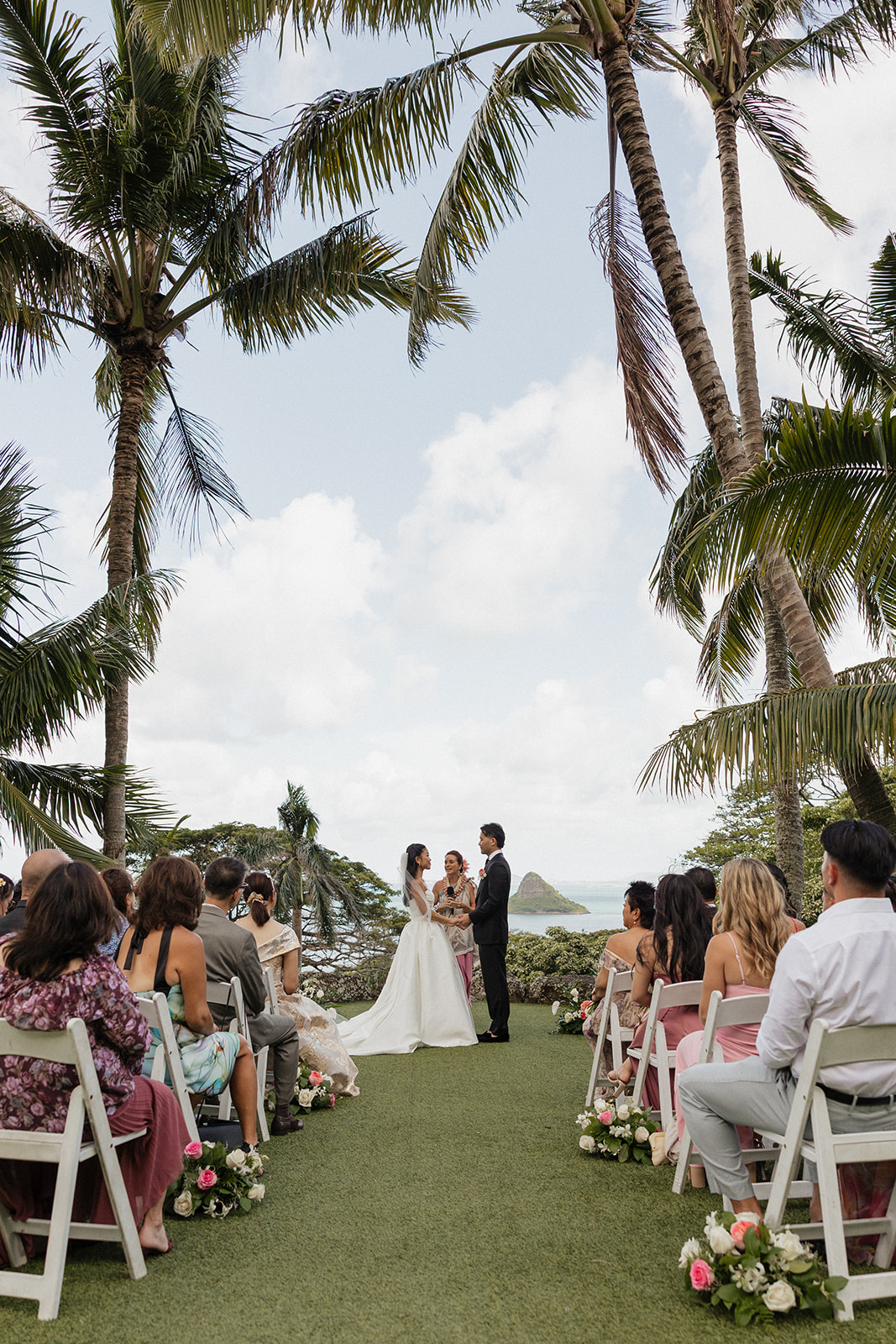 Wide ceremony shot taken from the aisle looking toward the altar, with guests seated in white chairs on either side framed by tall coconut palms, Mokoli'i Island floating on the horizon over Kaneohe Bay