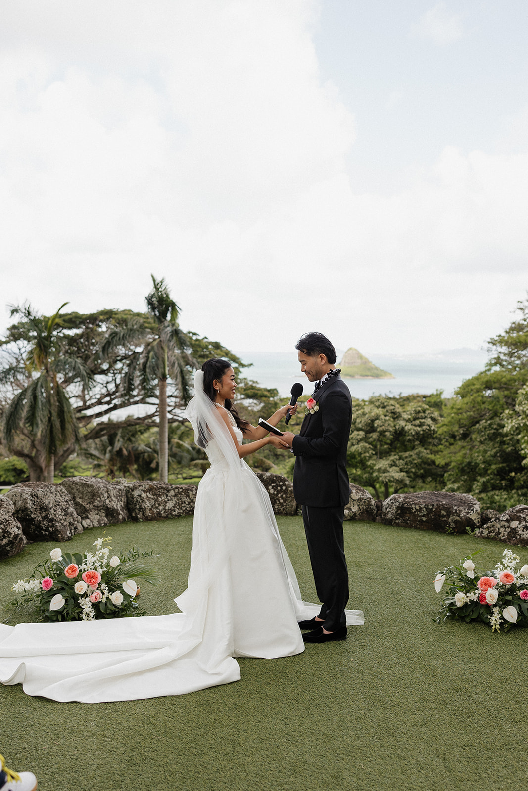 Bride and groom exchanging vows at their Kualoa Ranch Wedding ceremony, holding hands at the altar with floral arrangements at their feet, Mokoli'i Island and the ocean visible through the palm trees behind them