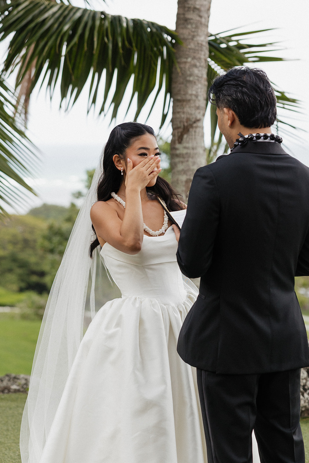 Bride wiping her tears during the outdoor ceremony while her groom reads his vows, wearing a strapless ball gown with a cathedral veil and a kukui nut lei under tropical palms