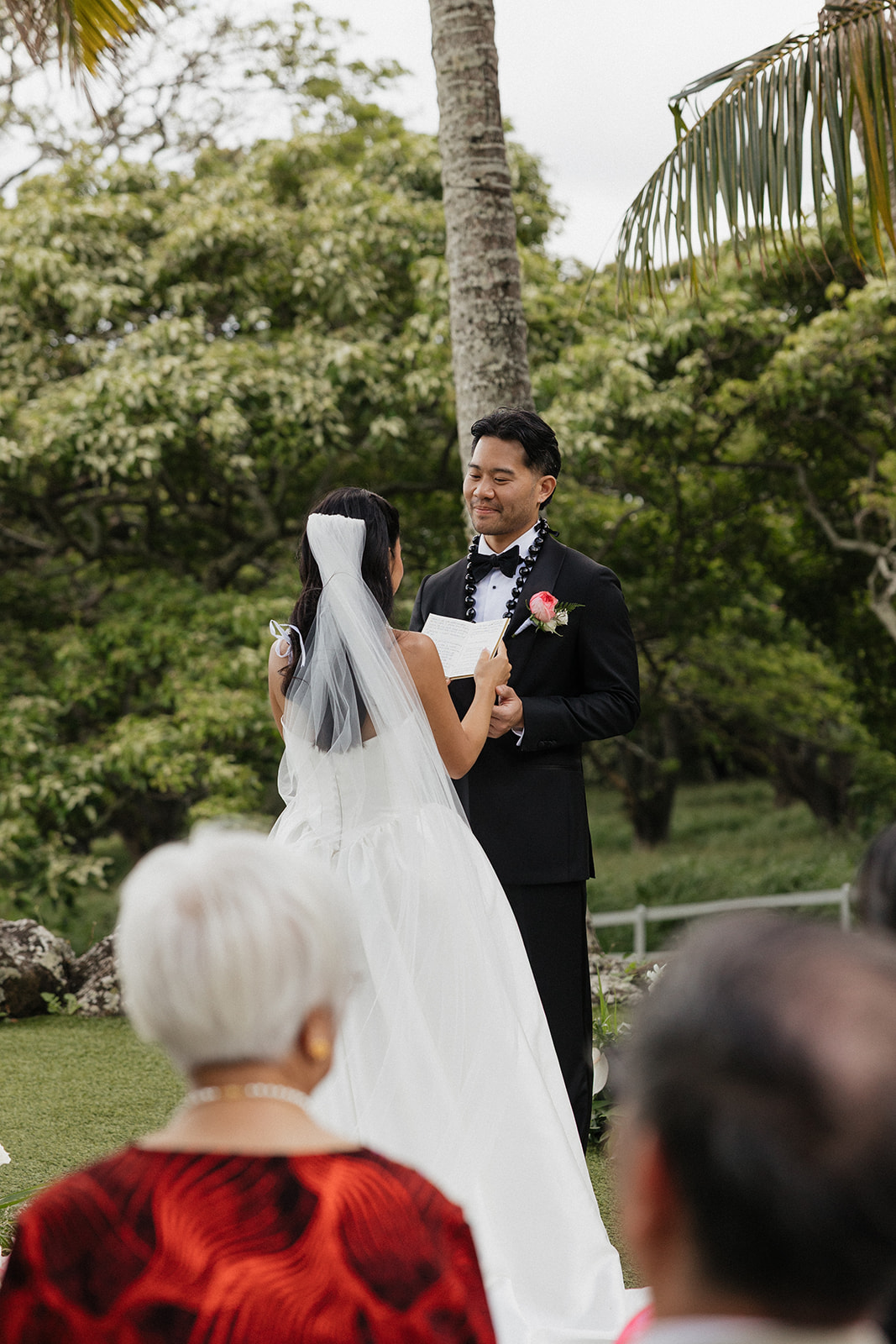 Groom reading his vows with an emotional smile during an outdoor wedding ceremony, wearing a black tuxedo and kukui nut lei against a lush tropical backdrop