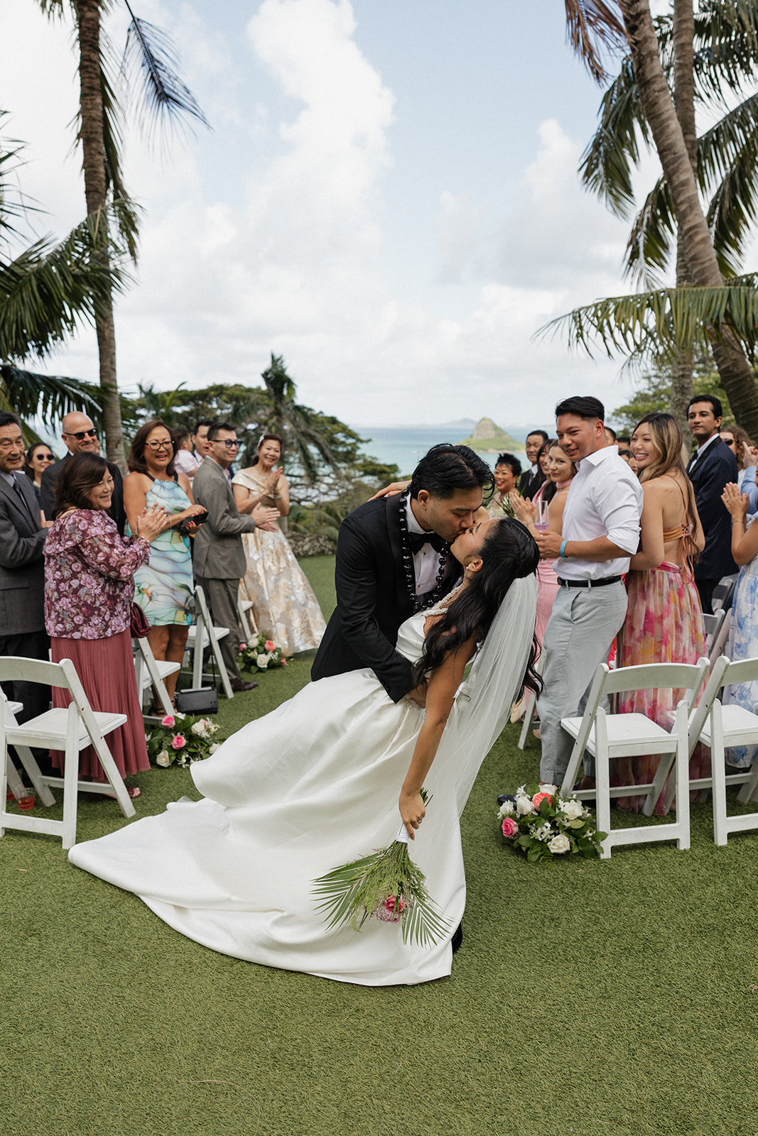 Groom dipping his bride for a kiss at their Kualoa Ranch Wedding recessional, with cheering guests lining the aisle and Mokoli'i Island visible over the ocean in the background