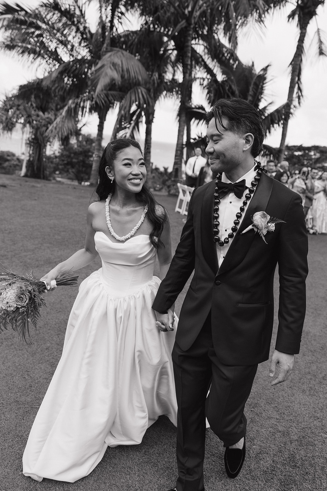 Black and white recessional photo of a beaming bride in a strapless ball gown and groom in a black tuxedo walking hand in hand after their ceremony, both wearing fresh flower lei under swaying palm trees