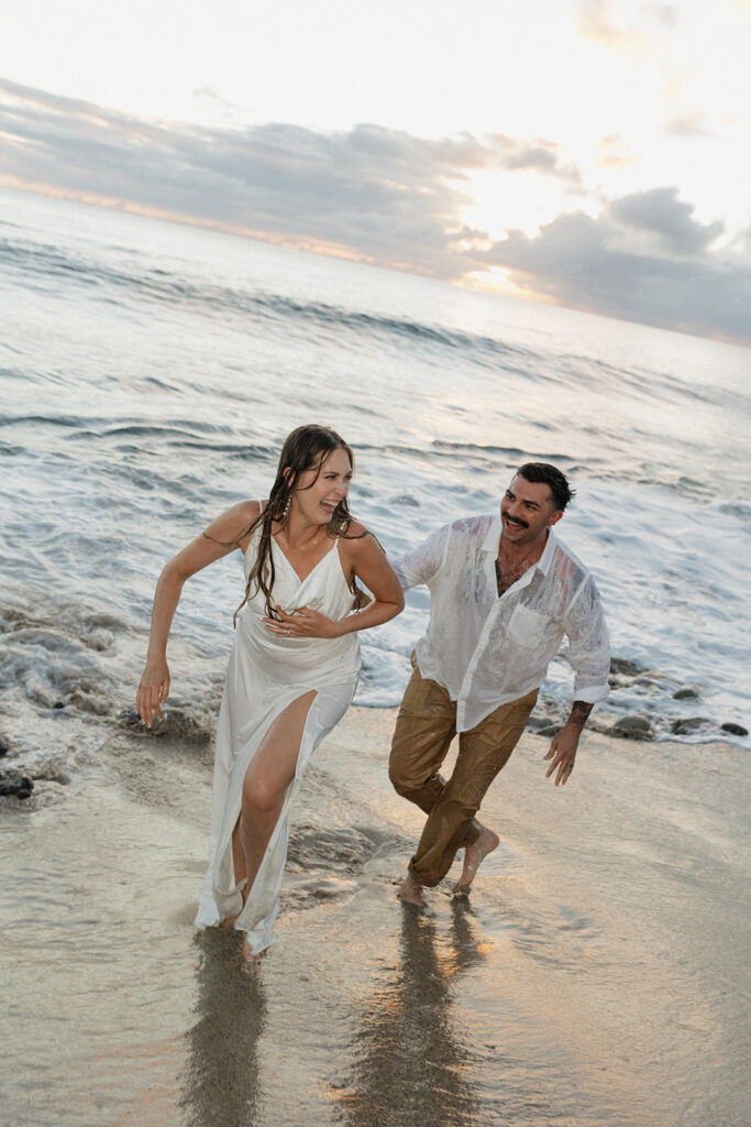 Bride and groom running out of the waves together, both laughing.