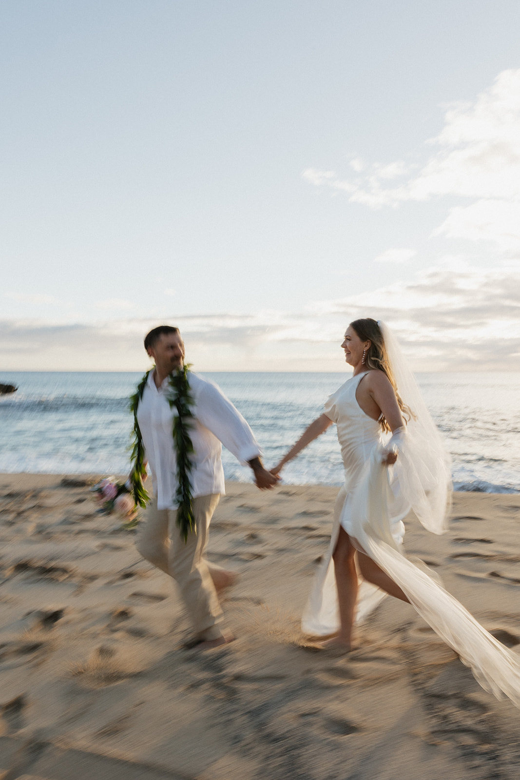 Bride and groom laughing as they walk hand in hand along the shore, her veil trailing behind.