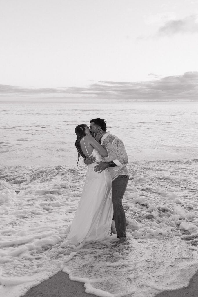 Black and white shot of the couple kissing while standing knee-deep in the ocean.