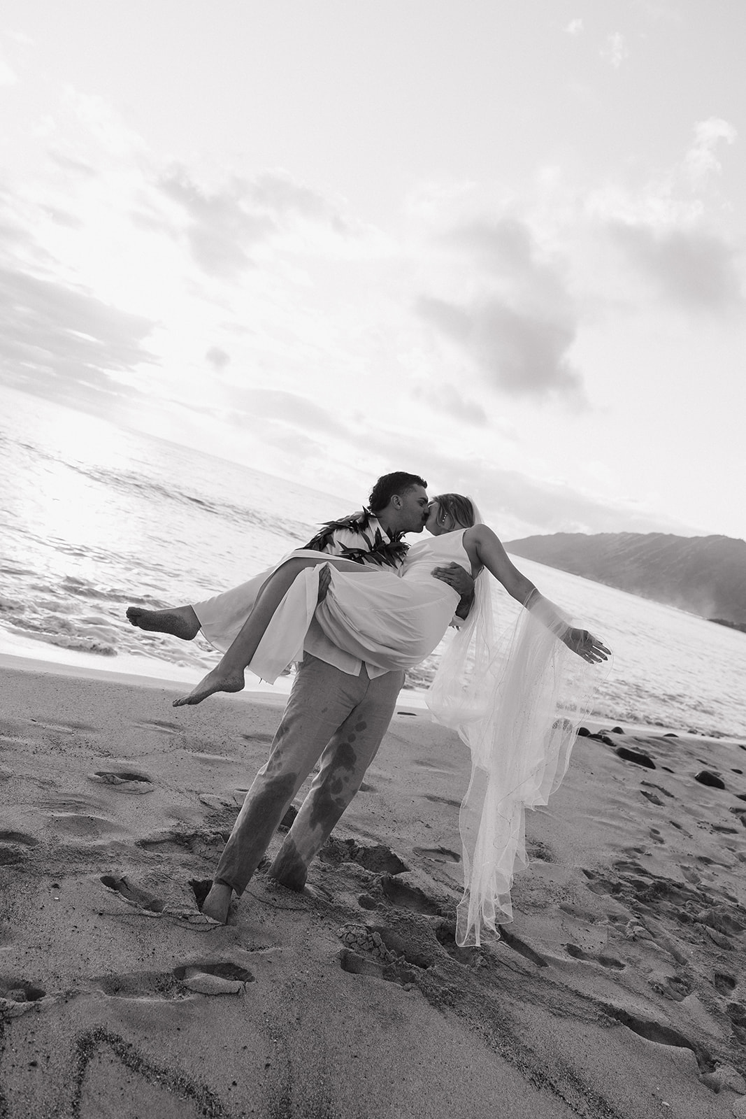 Black and white photo of the groom lifting his bride into the air for a kiss on the beach.