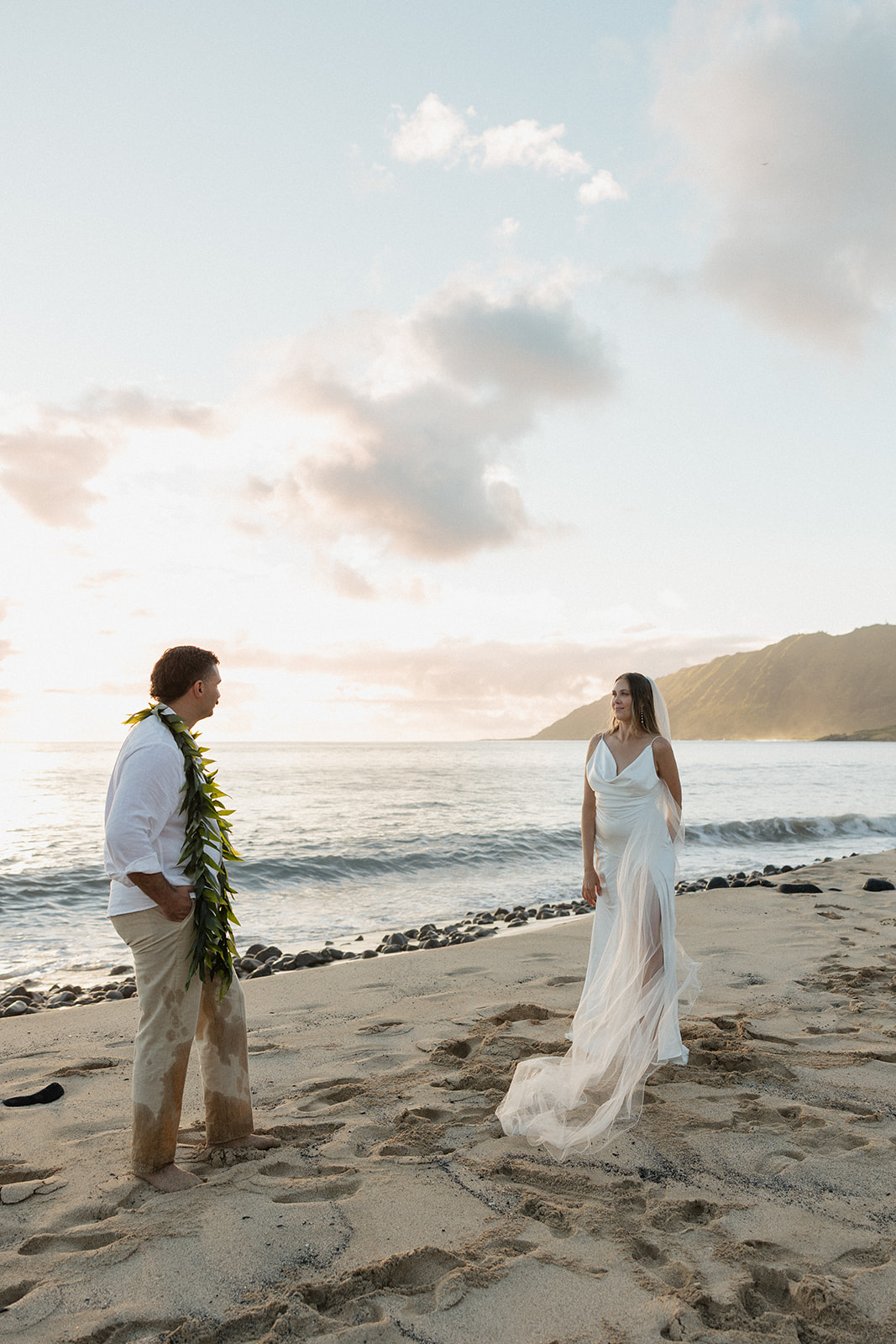 Bride and groom facing each other on the sand, her veil catching the breeze during their beach elopement.