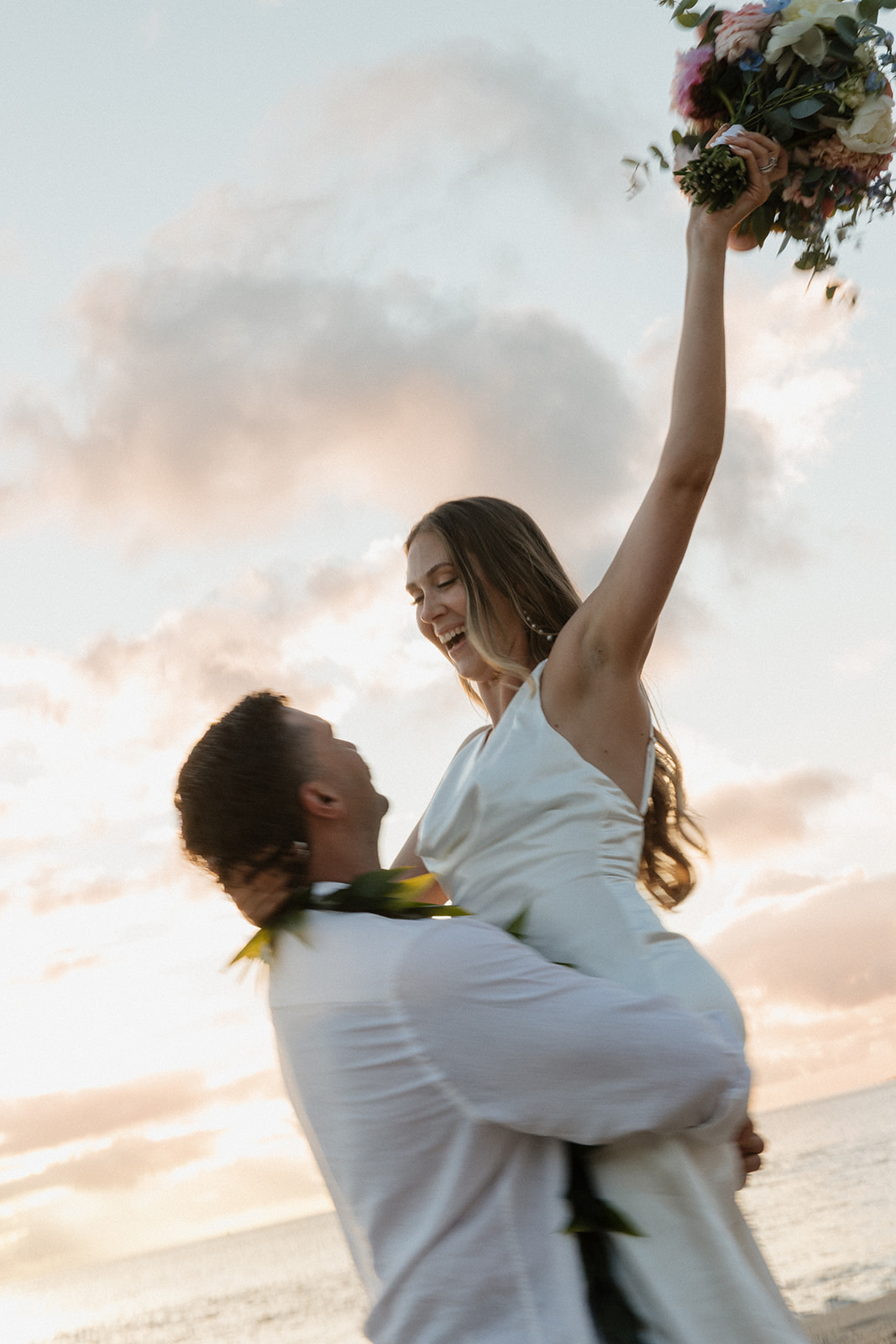 Groom lifting his bride into the air as she raises her bouquet, ocean sunset glowing behind them.