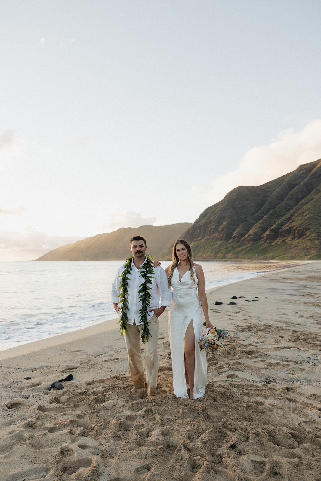 Bride and groom posing together on the sand with the mountains and coastline behind them.