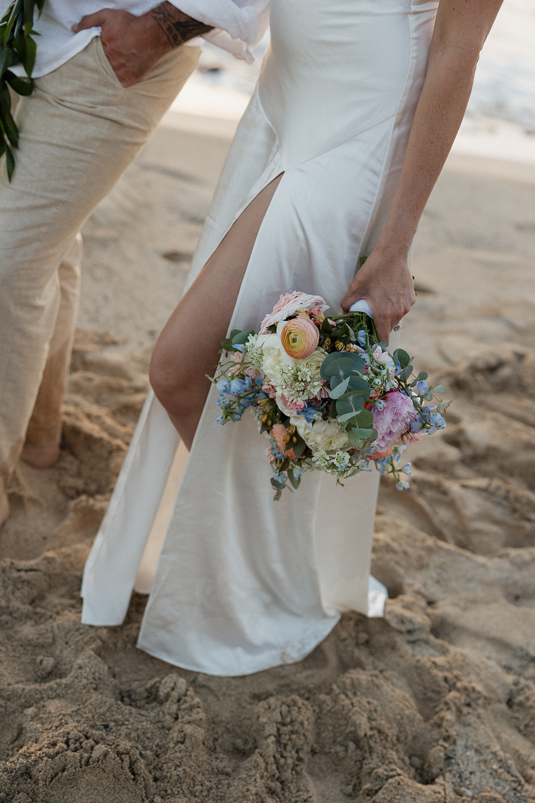 Close-up of the bride's bouquet against her dress slit as she stands next to her groom.