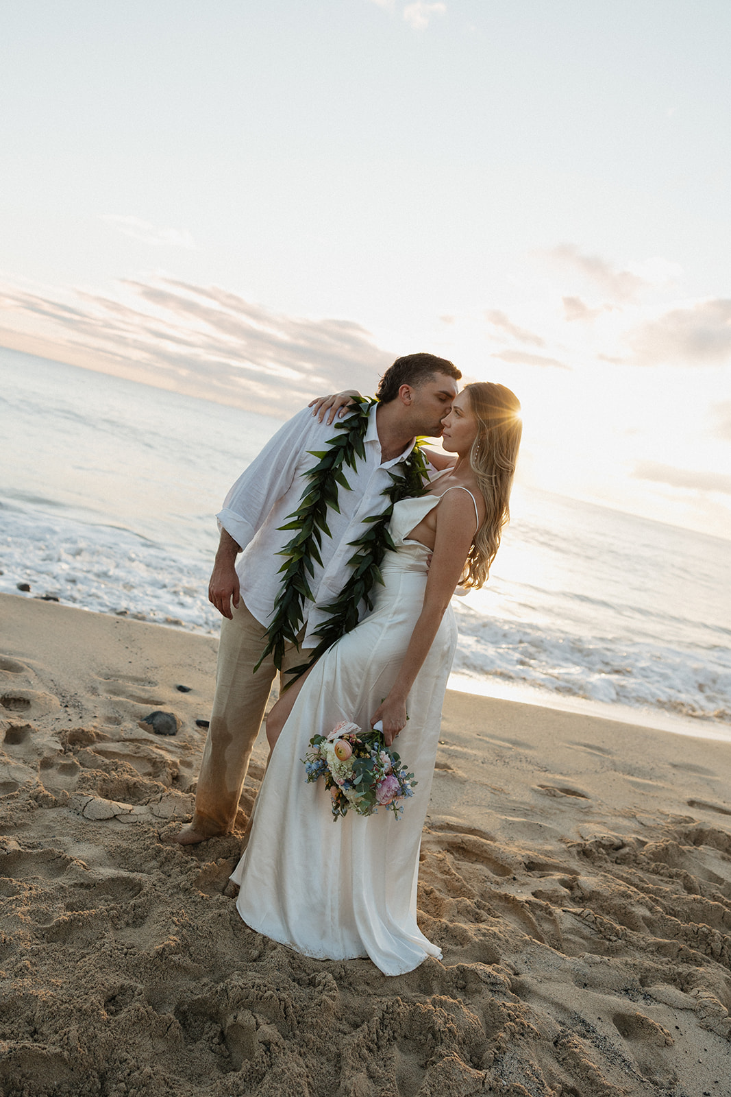 Groom kissing his bride on the beach as the sun sets behind them.