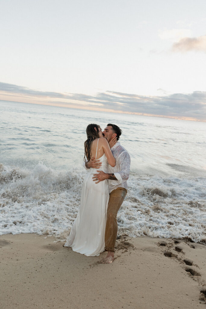 Newlyweds kissing in the waves, both soaked from head to toe.
