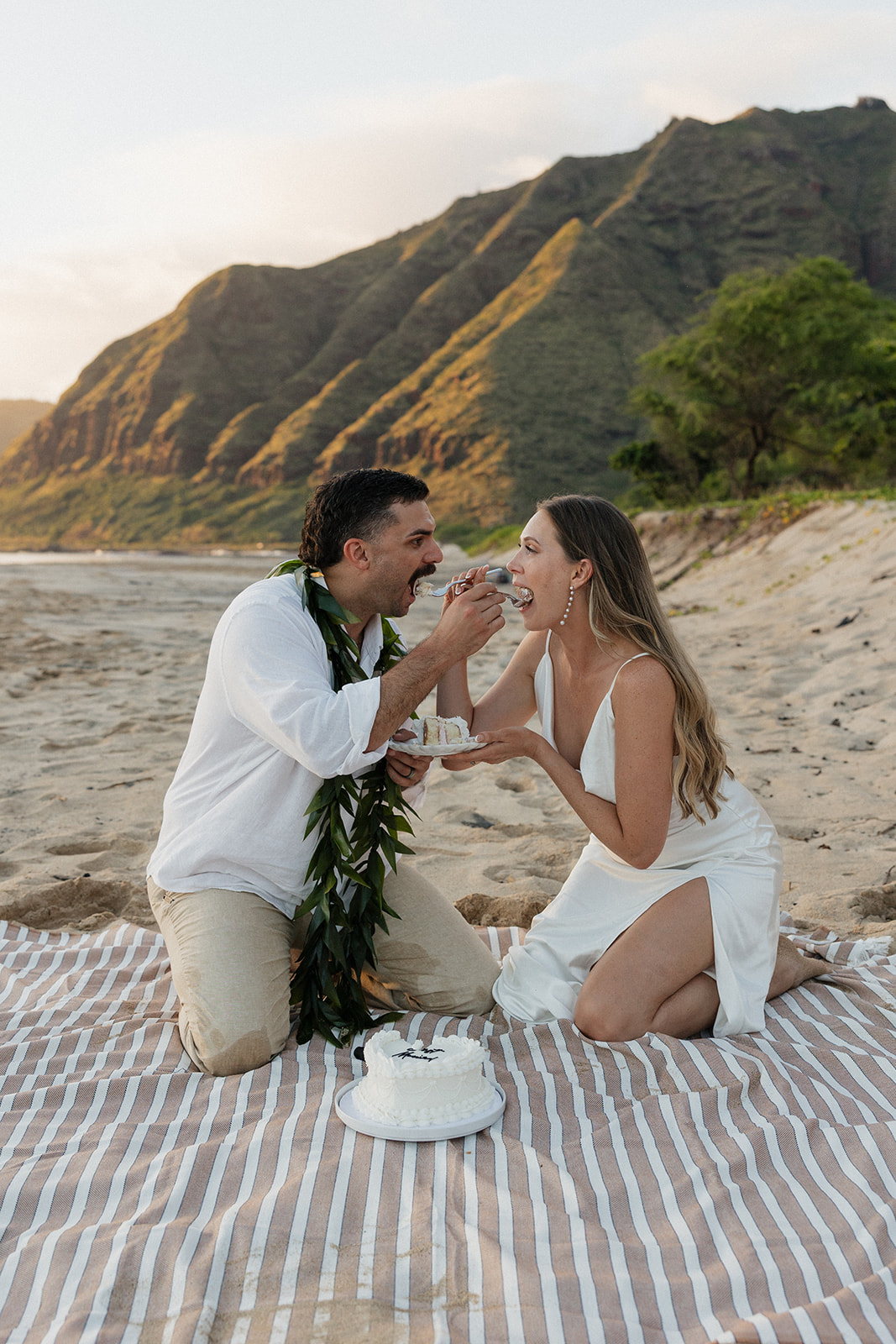 Groom feeding his bride a bite of cake on a striped picnic blanket in the sand.