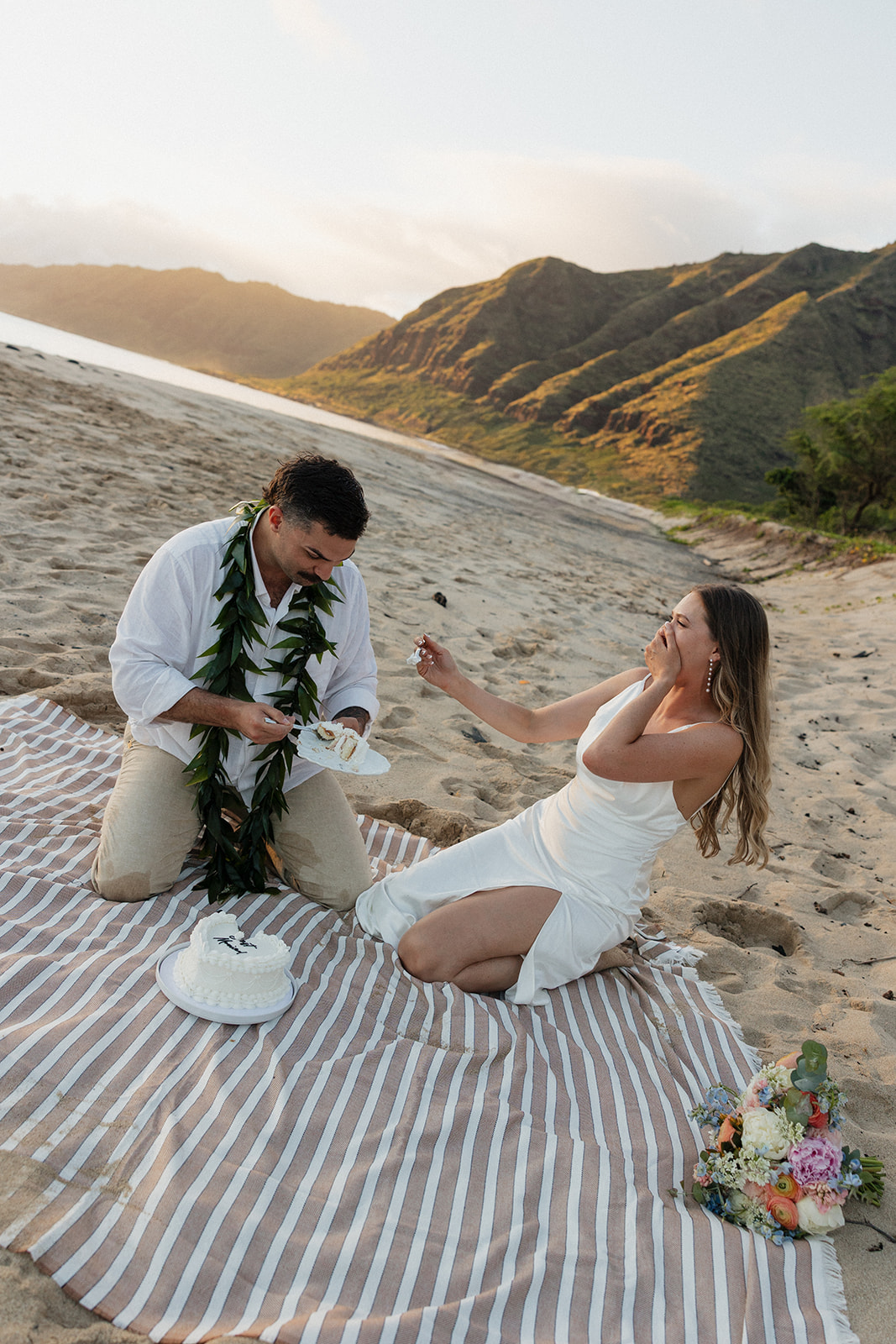 Bride and groom feeding each other cake on the beach at golden hour.