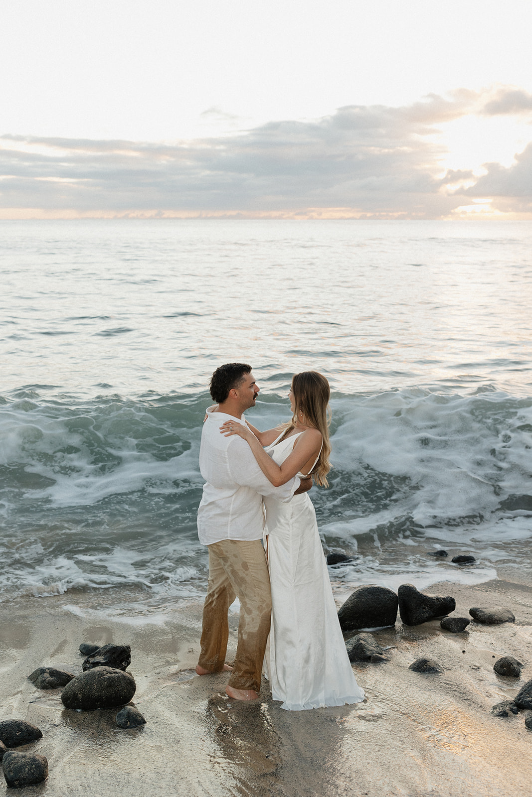 Bride and groom embracing in the shallow surf, waves crashing around them at sunset.