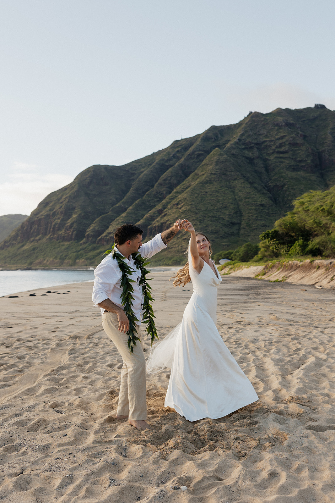 Groom twirling his bride in a joyful dance on the beach with mountains and ocean as the backdrop.