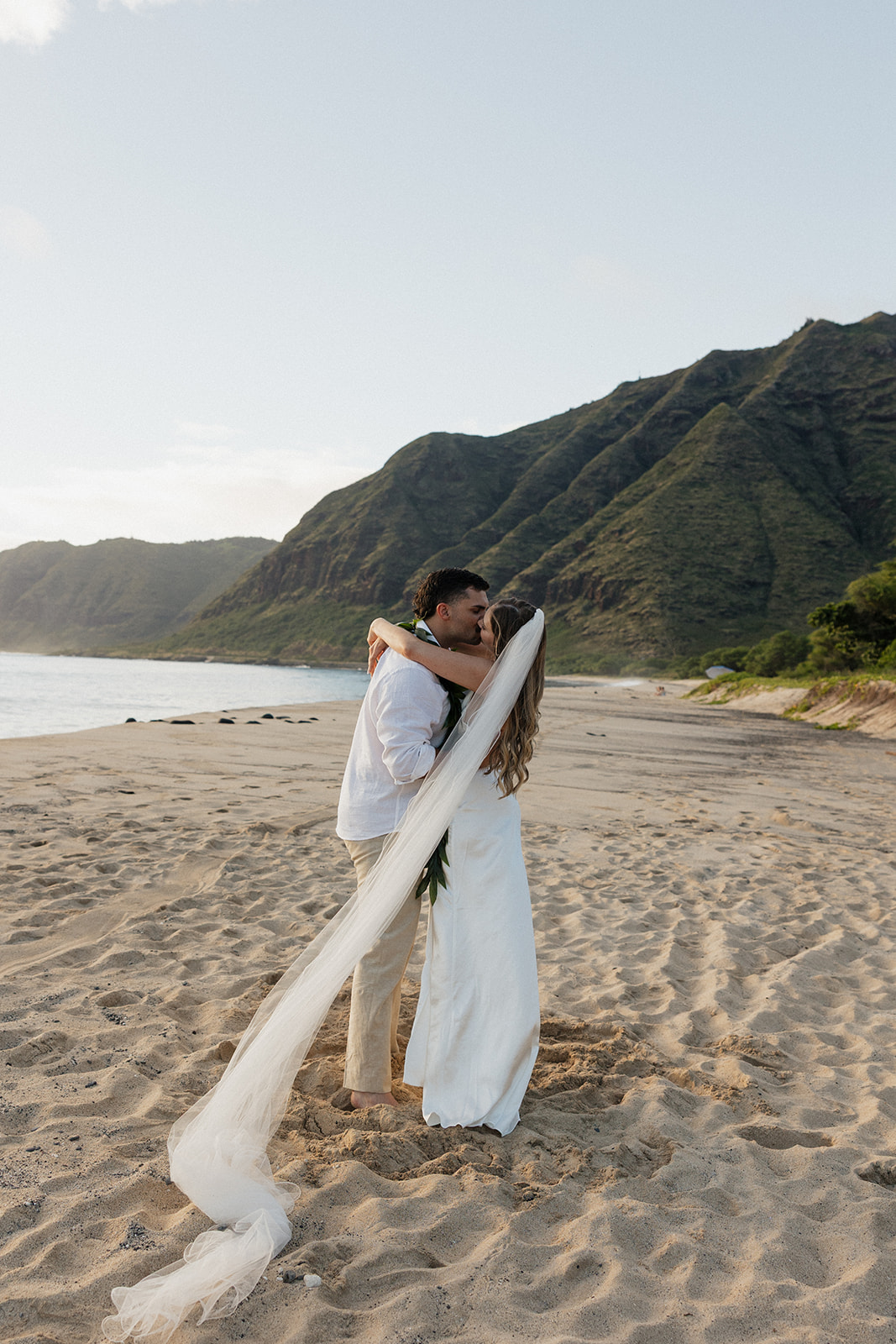 Bride and groom kissing on the beach, her veil trailing out behind them in the wind.