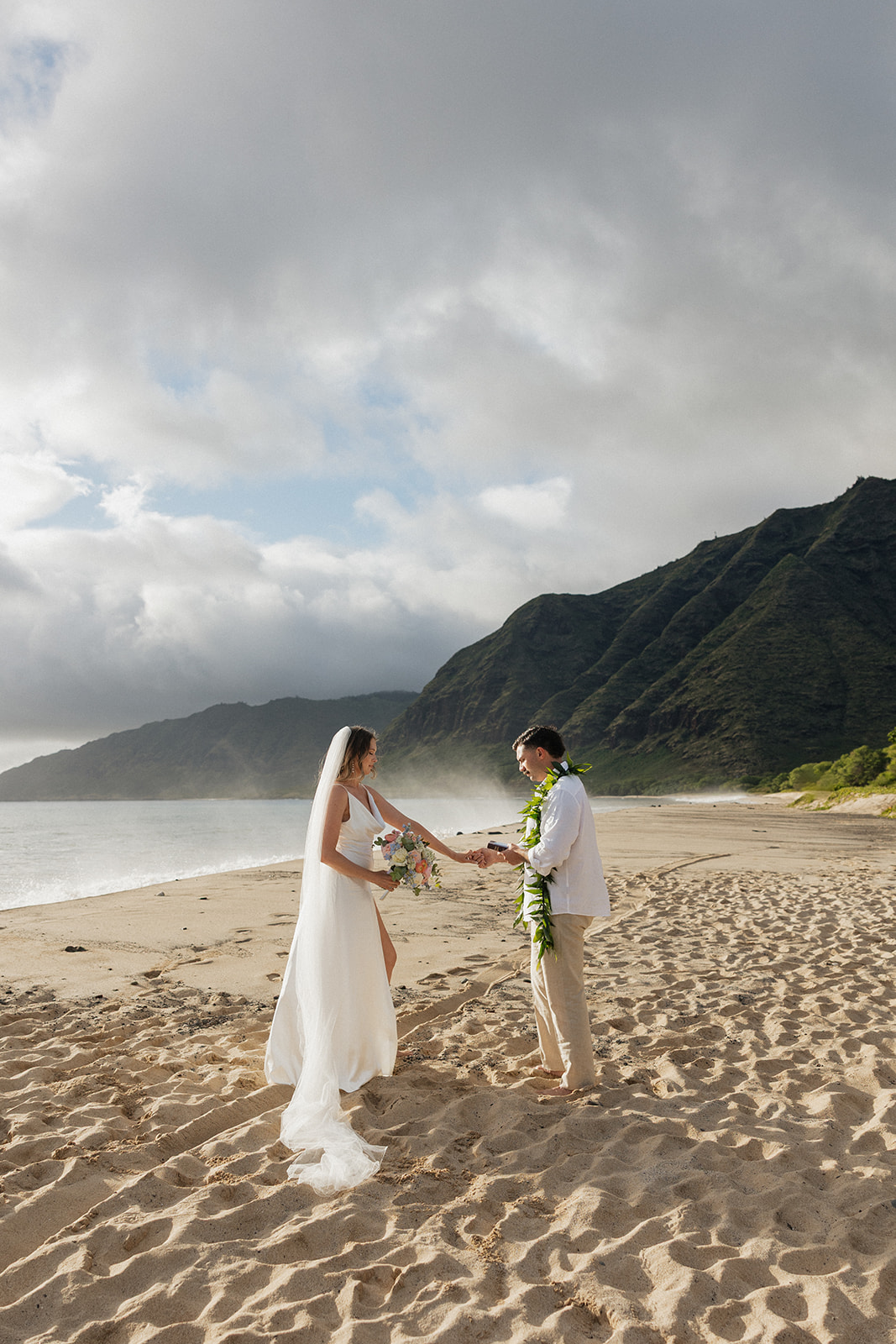 Bride and groom holding hands during their vow exchange, waves rolling in beside a dramatic Oahu mountain backdrop.