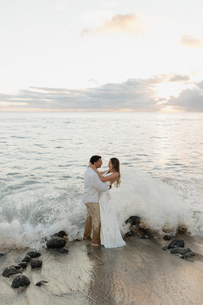 A wave crashes behind the bride and groom as they hug on the rocky shore.