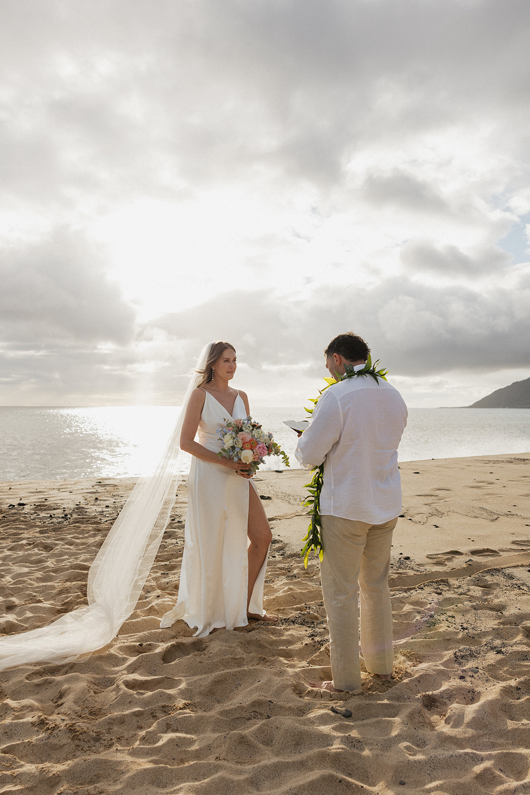 Bride holding her bouquet as she listens to the groom's vows with the ocean glowing behind them.