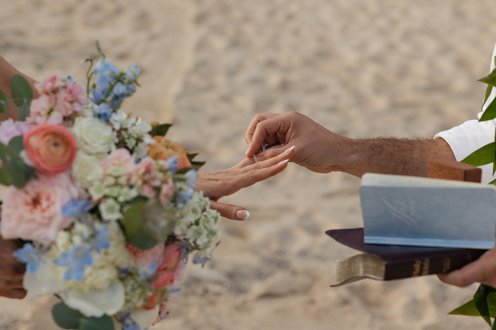 Close-up of the groom placing a wedding ring on the bride's finger during their beach elopement ceremony, bouquet in the foreground.
