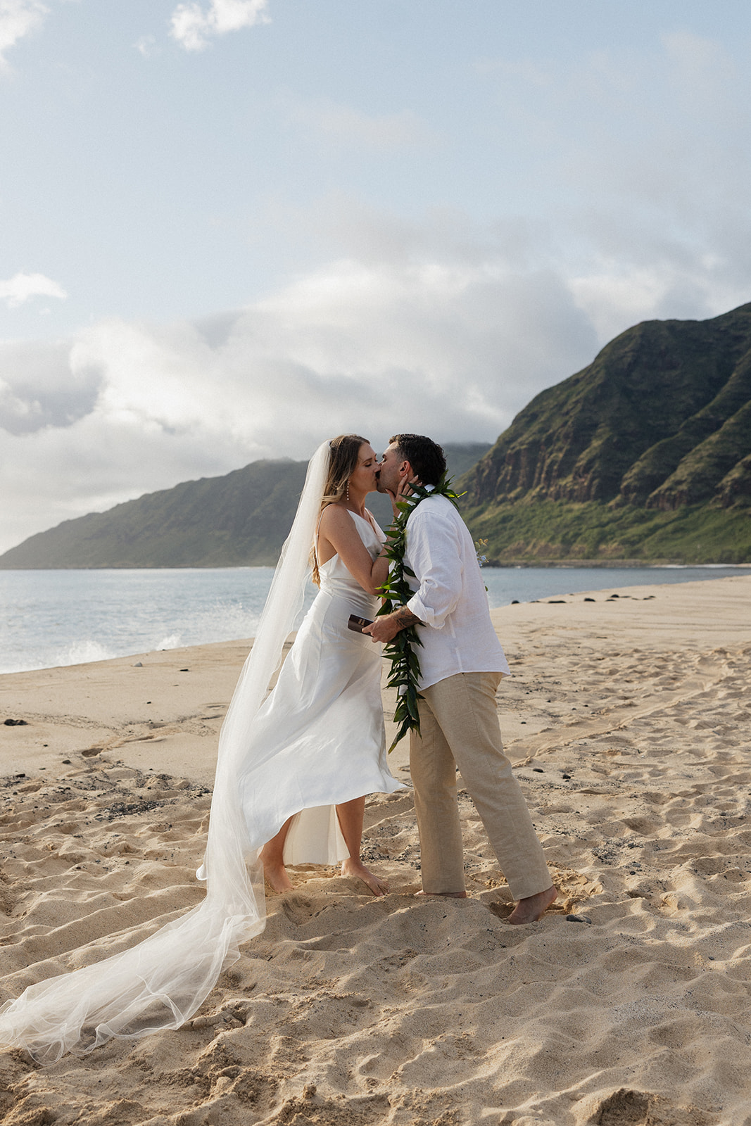 Bride and groom sharing a kiss on the sand during their Oahu beach elopement, with green mountains and the beach behind them.