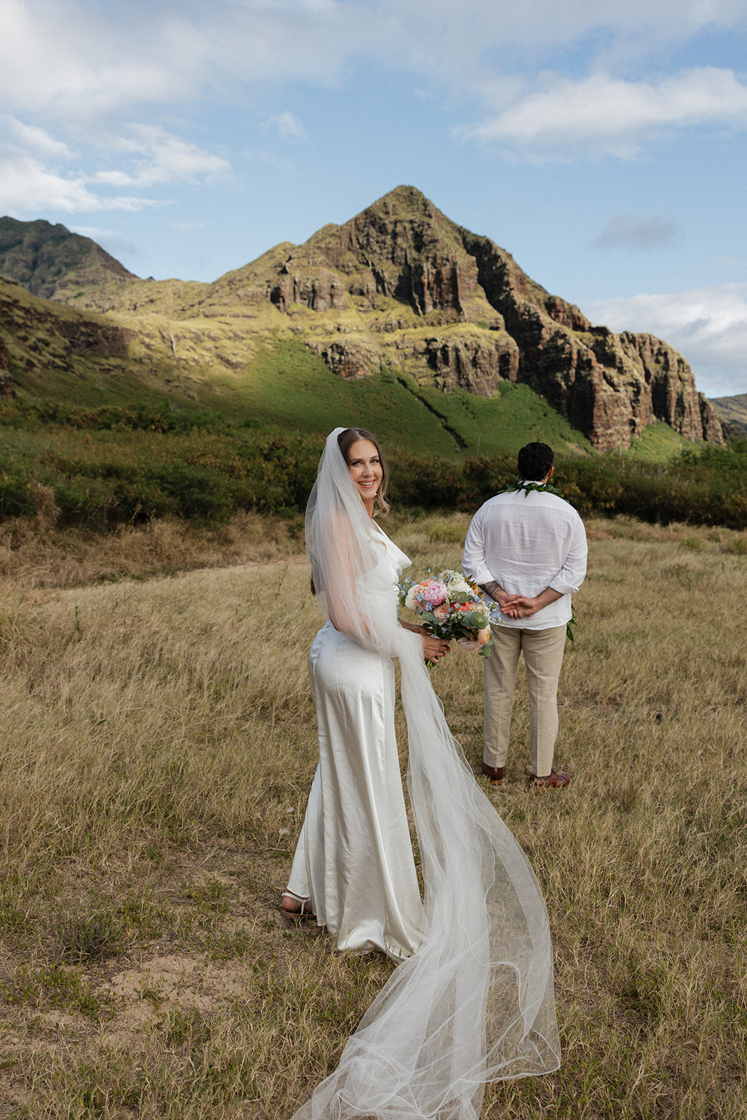 Bride smiling back at the camera while walking through tall grass toward her groom, mountain peak in view.