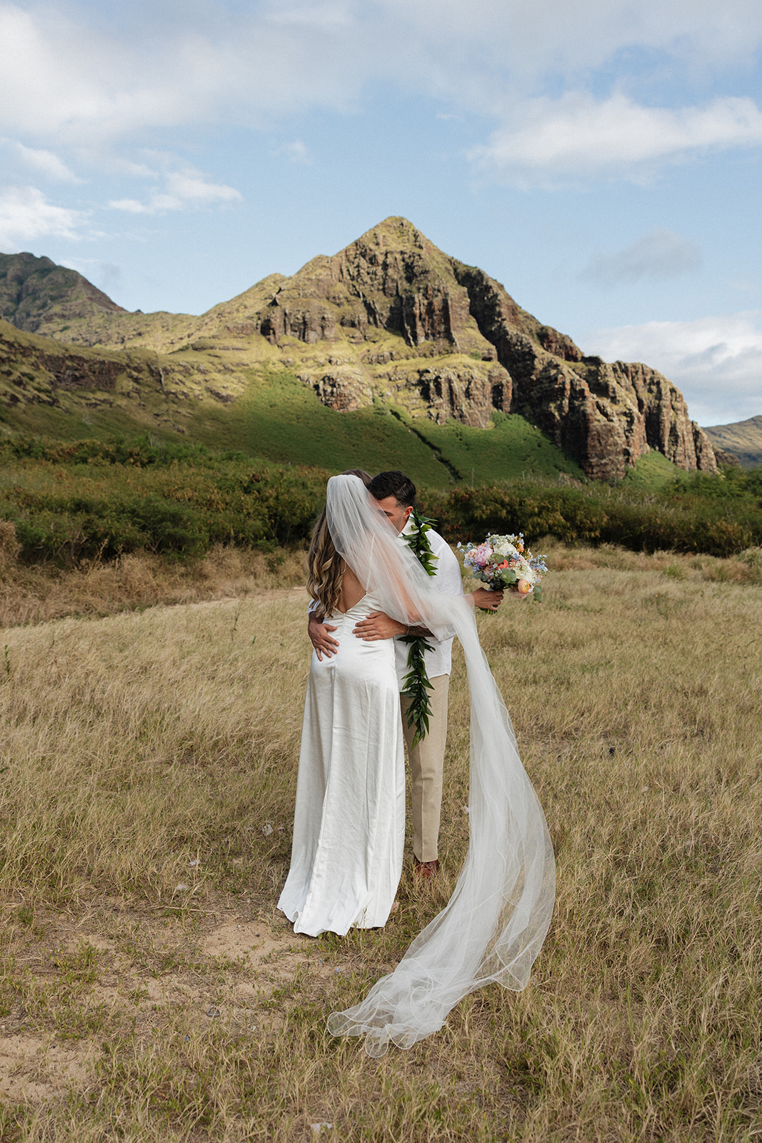 Groom embracing his bride in a grassy field with a dramatic mountain peak towering behind them.