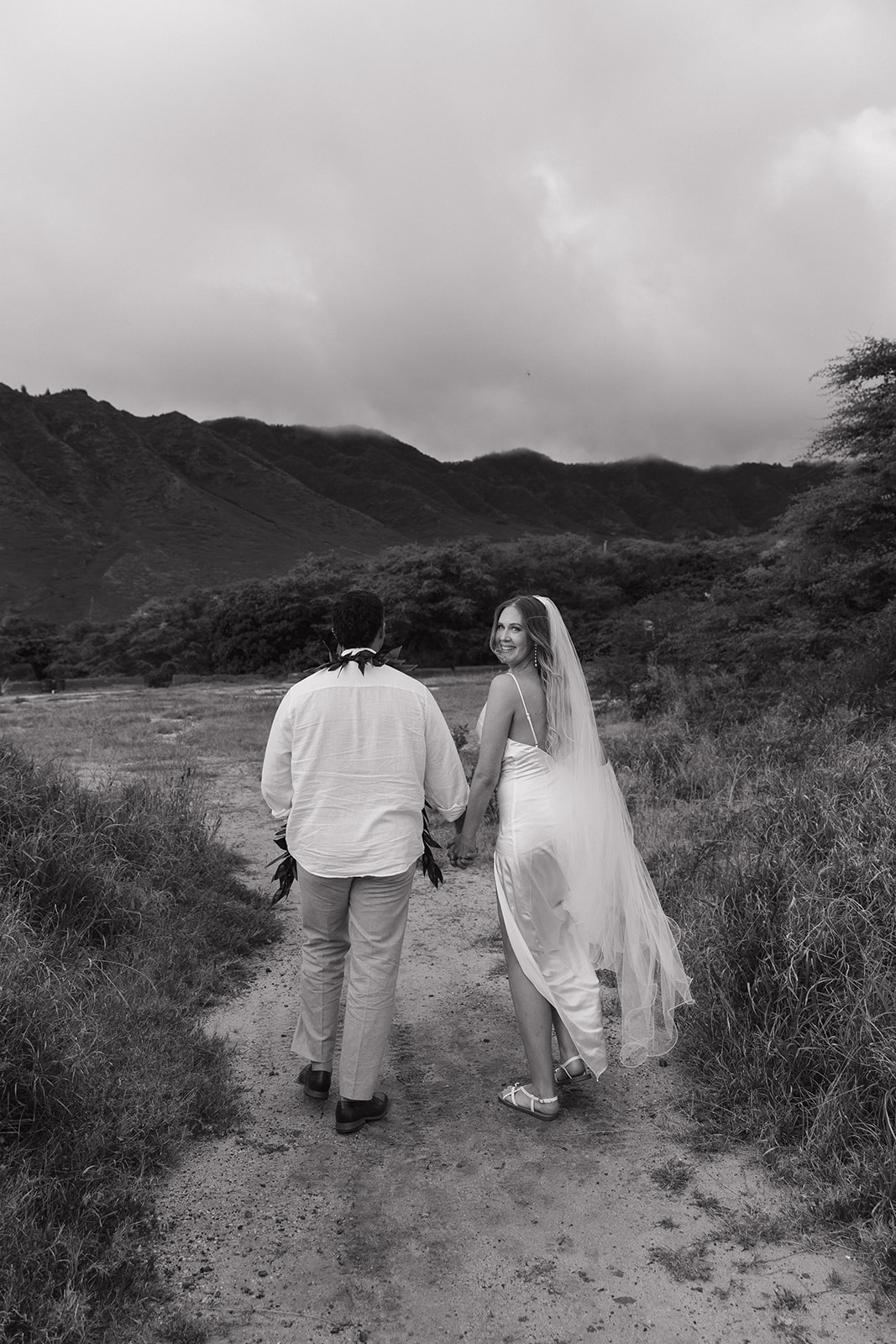 Bride glancing back with a smile as the couple walks together along a grassy trail near the coast.