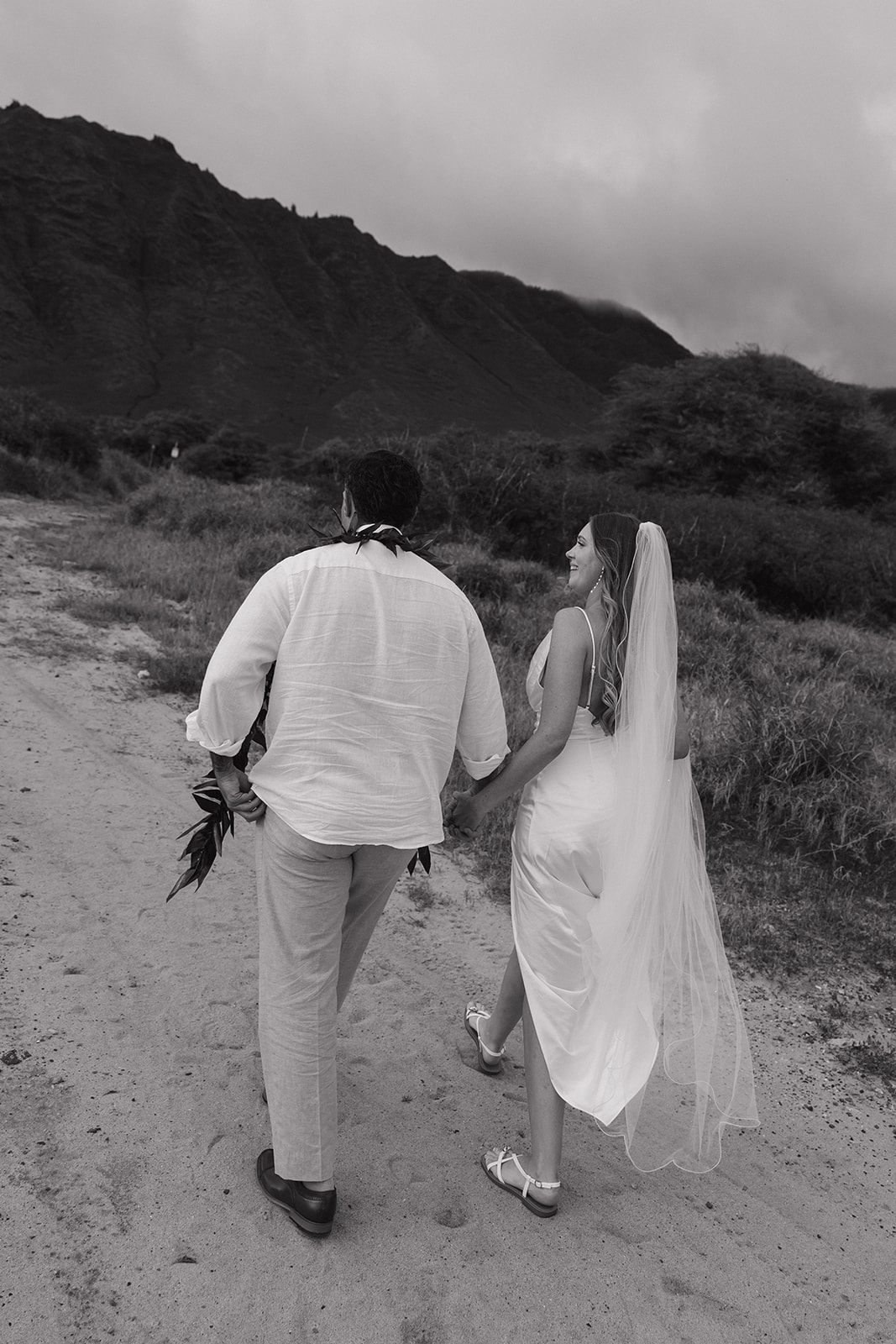 Black and white photo of the newlyweds walking hand in hand down a dirt path after their beach elopement.