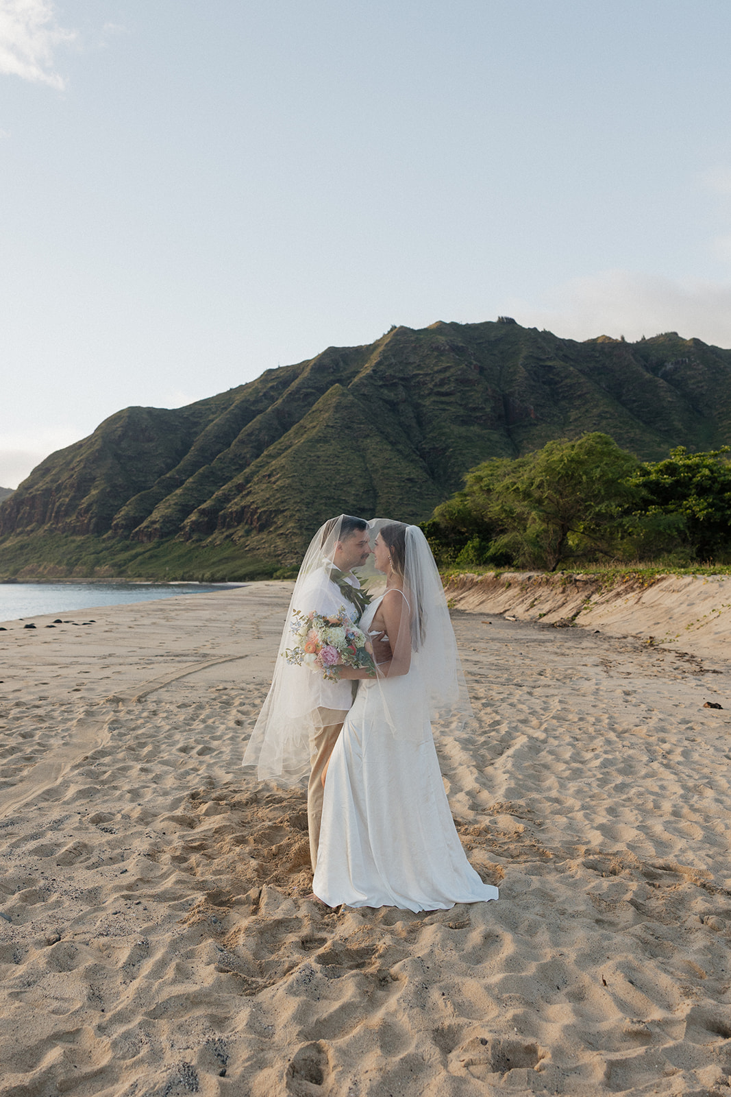 Bride and groom sharing a moment under her veil on the beach, mountains behind them.