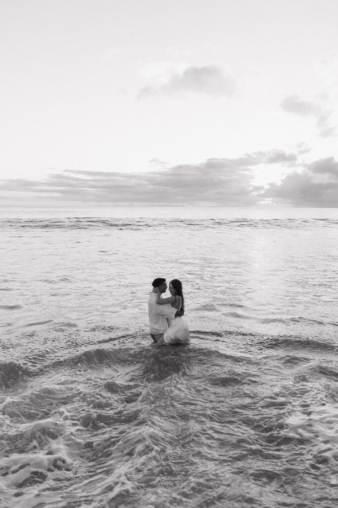 Black and white photo of the couple embracing far out in the water at sunset.