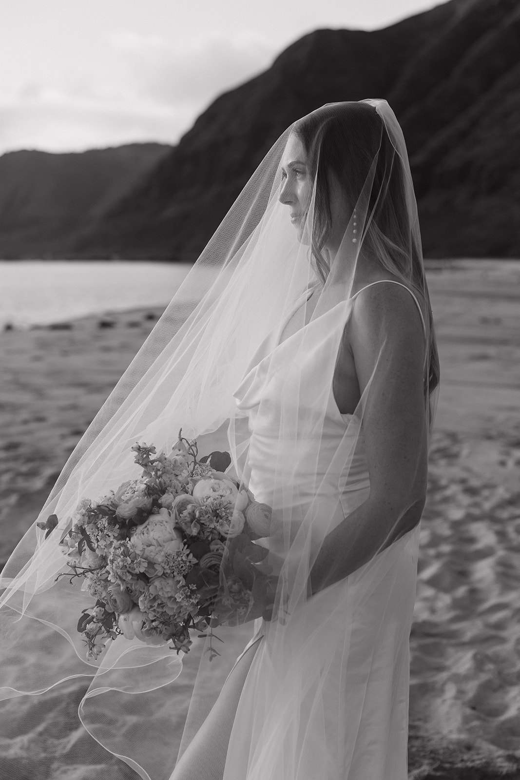 Black and white portrait of the bride in her veil, holding her bouquet by the water.