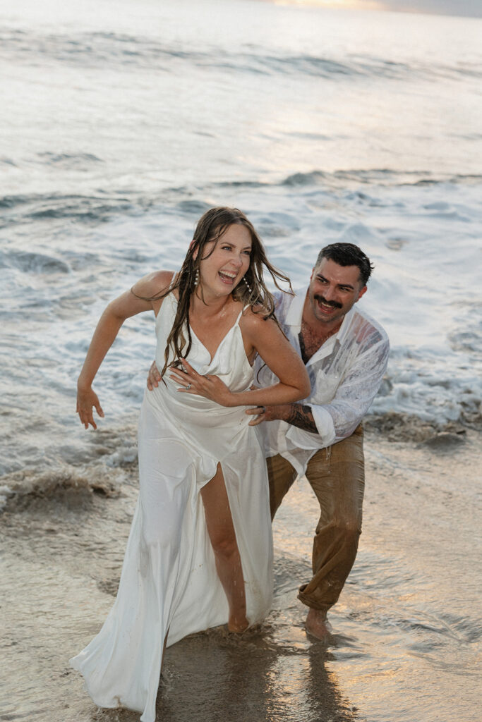 Bride laughing as her groom chases her out of the surf, dress completely drenched.