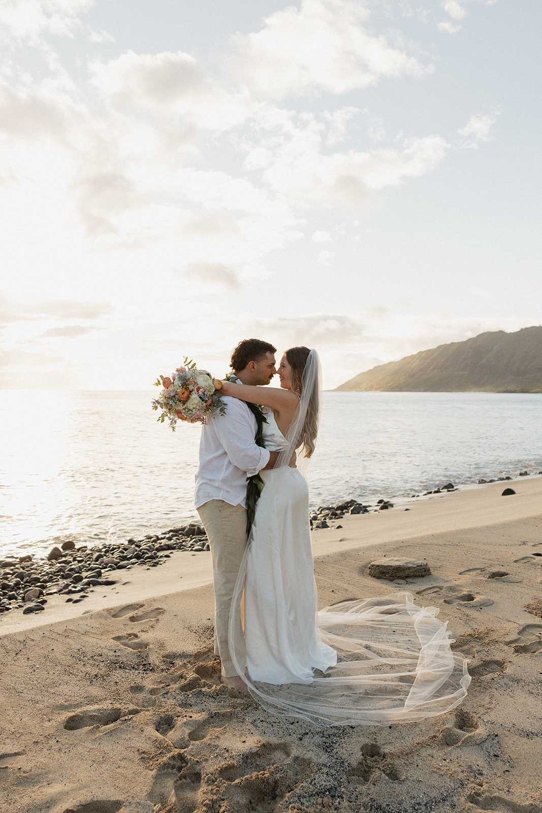 Bride and groom embracing on a rocky stretch of beach as the sun sets over the water.