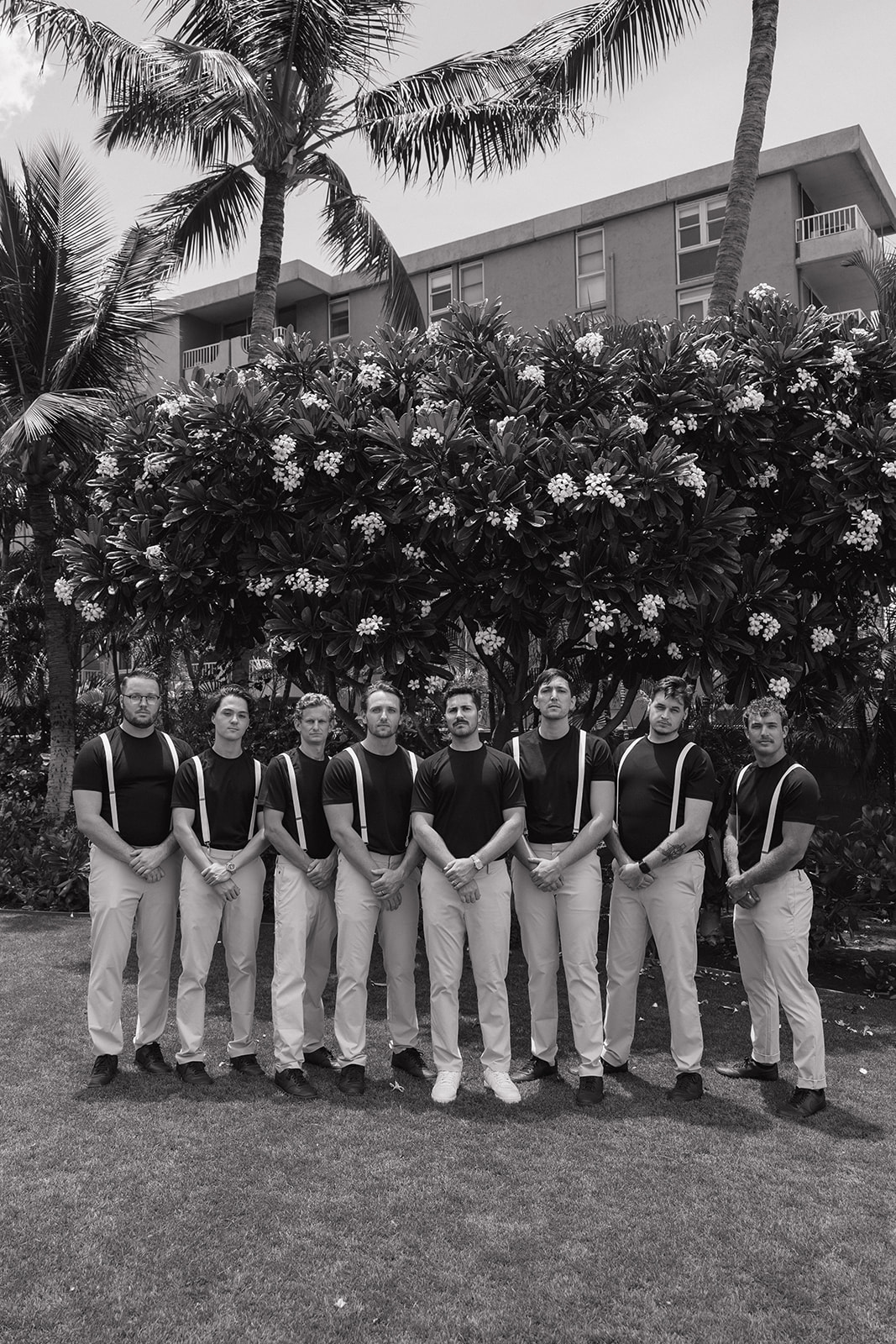 Black and white portrait of the groomsmen lined up in matching suspenders in front of a flowering plumeria hedge.