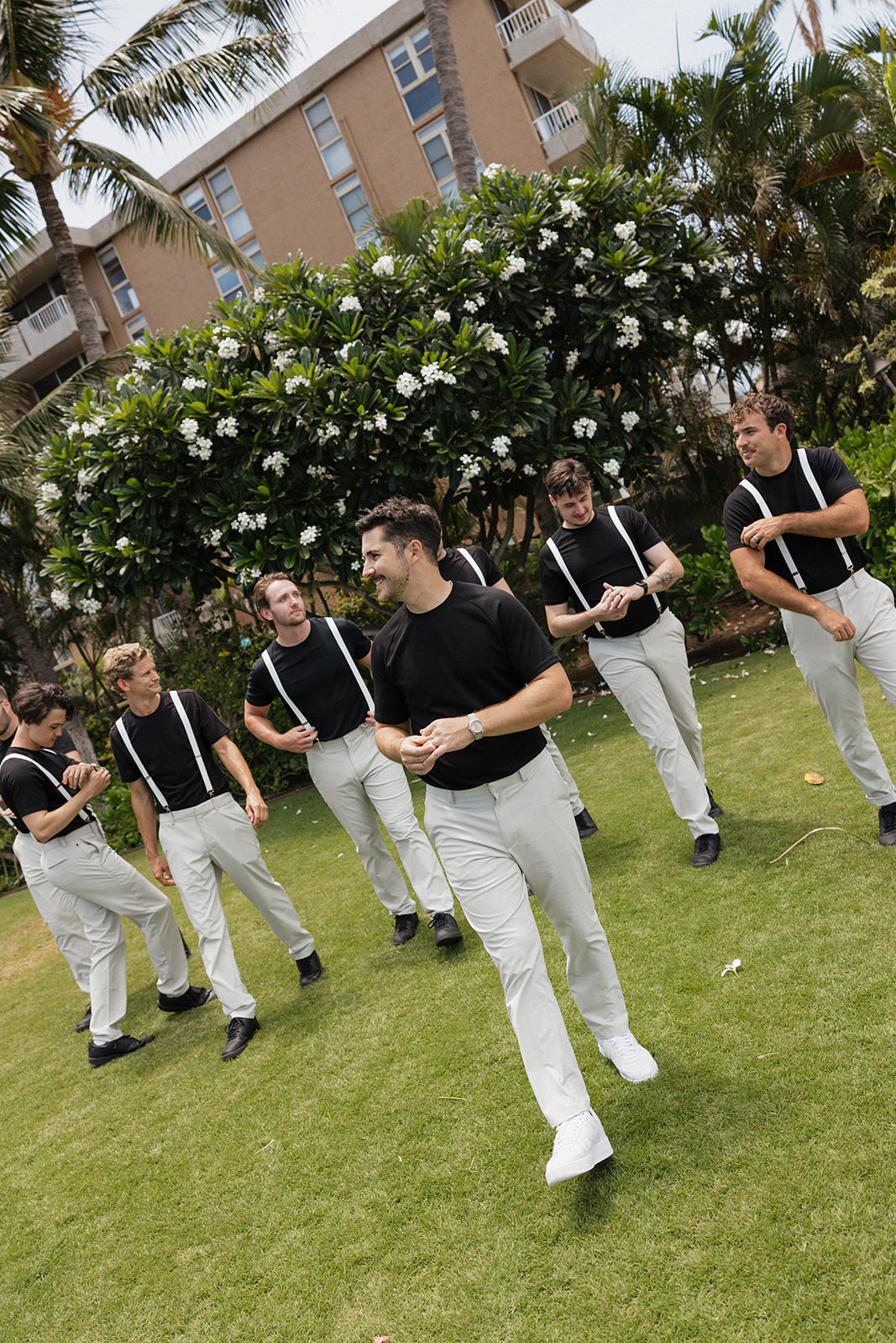 Groomsmen in black tees and suspenders walk and laugh together across the lawn before the ceremony.