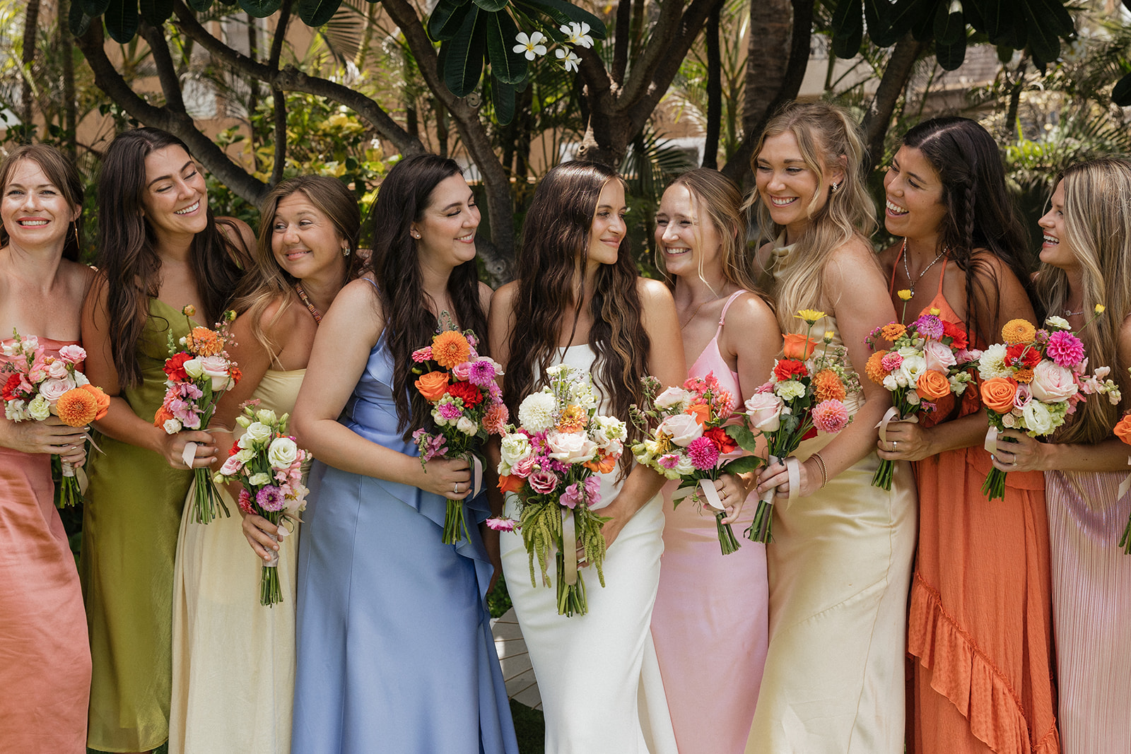 Bridesmaids in a rainbow of dresses gather around the bride, laughing together while holding their bouquets at this Maui wedding venue.