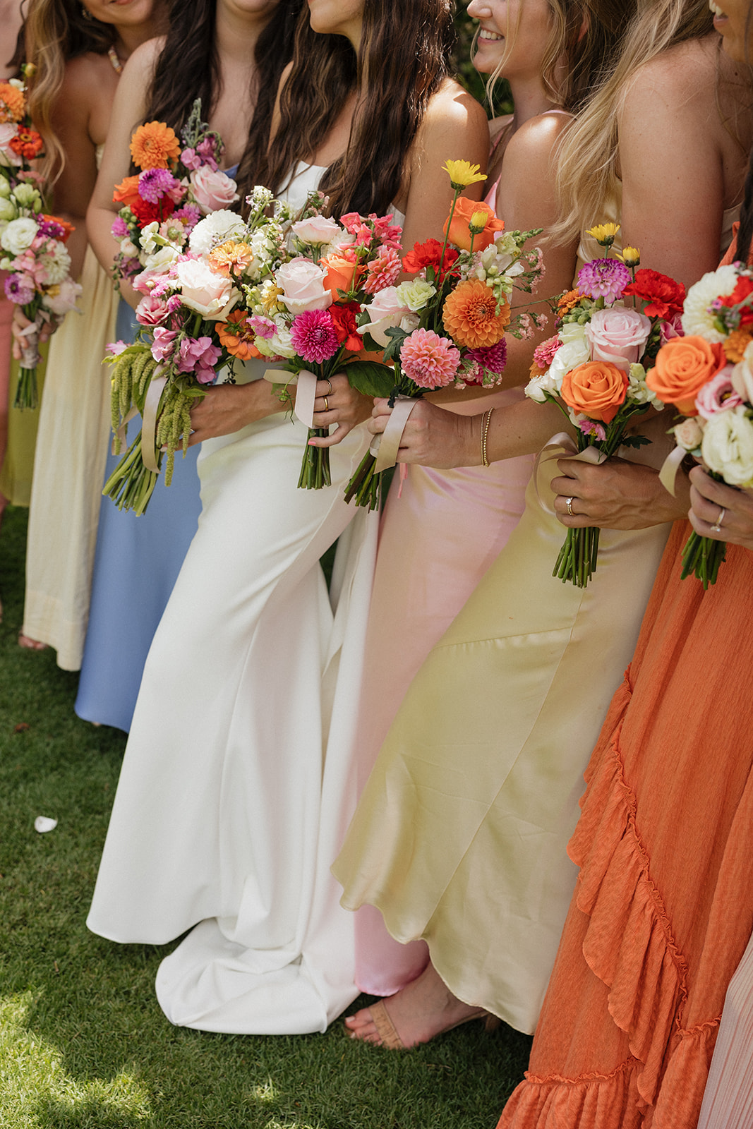 Bridesmaids in a colorful row of pastel and jewel-toned dresses hold vibrant bouquets of roses and dahlias.