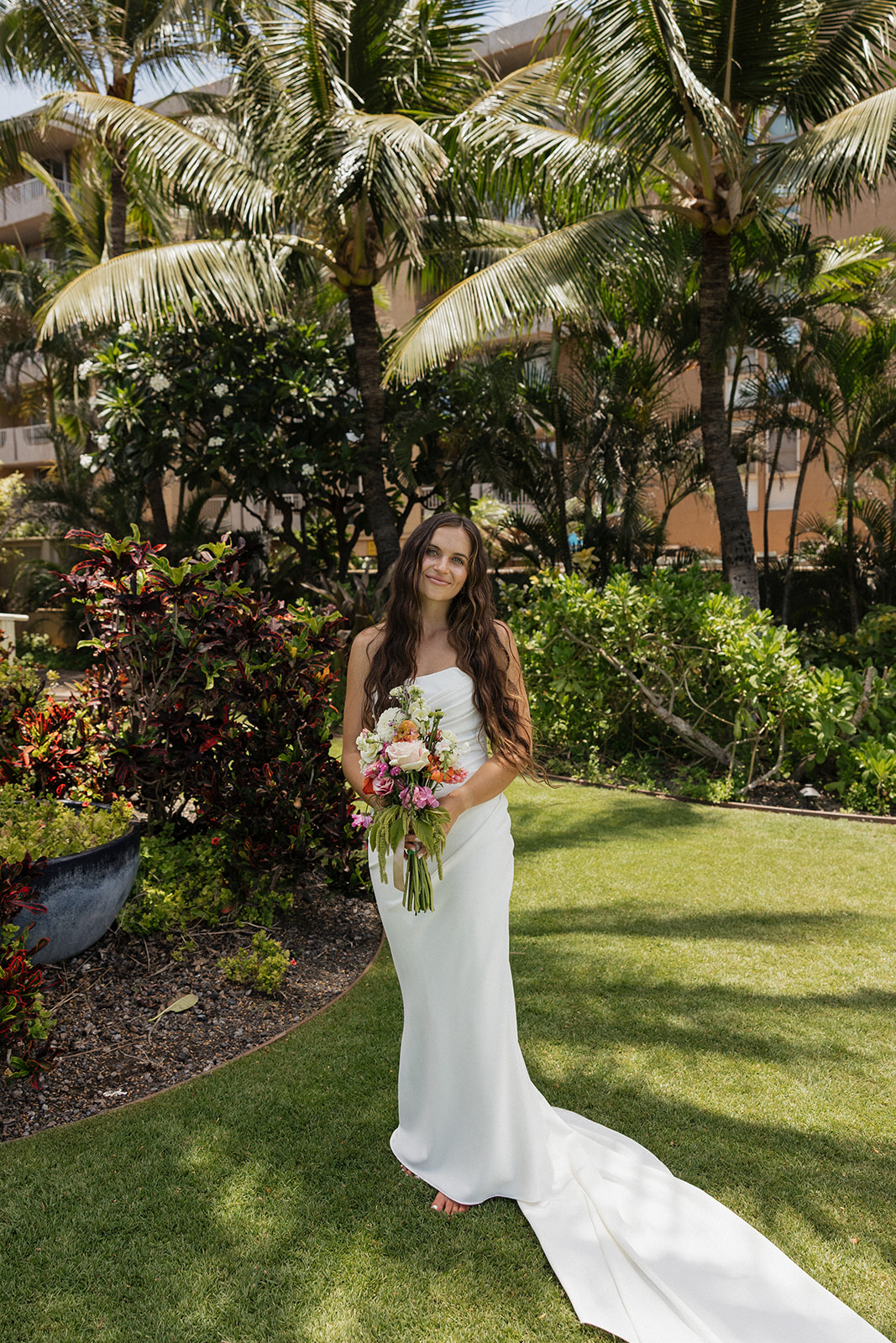 Bride poses among lush tropical greenery and palm trees, holding her bouquet and smiling at the camera.