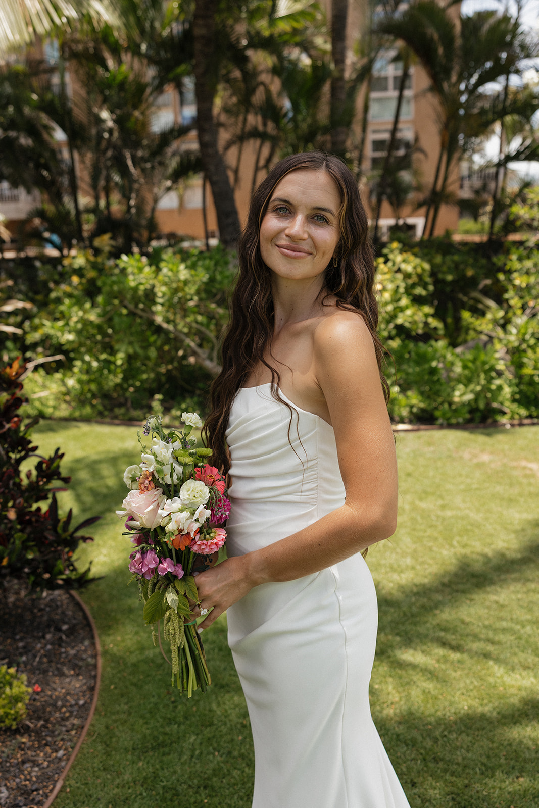 Bride in a strapless gown smiles softly while holding her colorful bouquet in the tropical garden.
