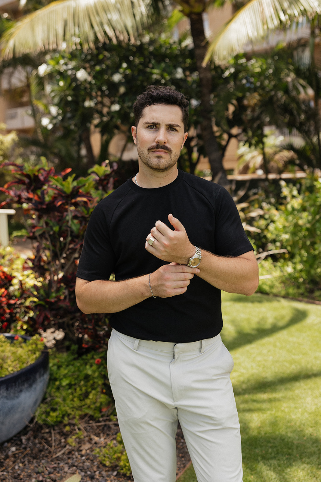 Groom portrait in a black tee and light trousers standing among tropical greenery before the ceremony.